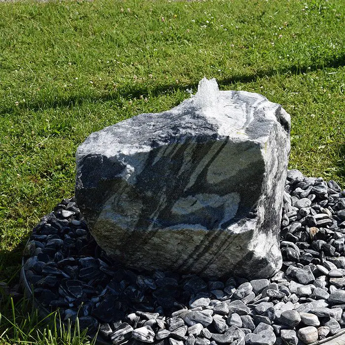Grey and white veined marble boulder fountain with water emerging at the top, on dark pebbles surrounded by lawn